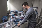A person wearing safety equipment operates a mitre saw to cut a metal piece.