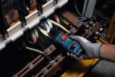 A person wearing safety equipment uses a fork meter to test electrical wires in a panel.