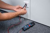 Person testing a wall socket voltage with a digital clamp meter on a carpeted floor.