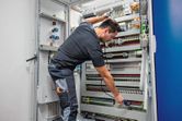 Person wearing safety equipment tests an electrical control panel with a voltage tester.