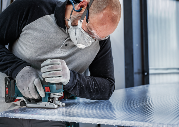 Person wearing safety equipment uses a jigsaw to cut a sheet of corrugated plastic.