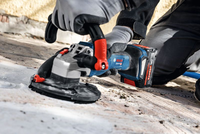 A person wearing safety equipment uses a cordless concrete grinder on a floor surface.