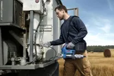 A person wearing safety equipment uses a cordless grease gun to service farm machinery.