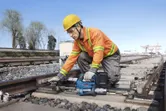 A person wearing safety equipment uses a cordless grease gun to maintain railway tracks.