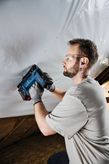 Person wearing safety equipment uses a cordless tool to fasten material to a ceiling.