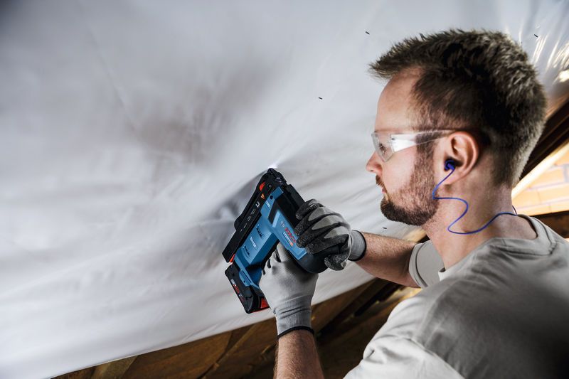 Person wearing safety equipment uses a cordless tool to fasten material to a ceiling.