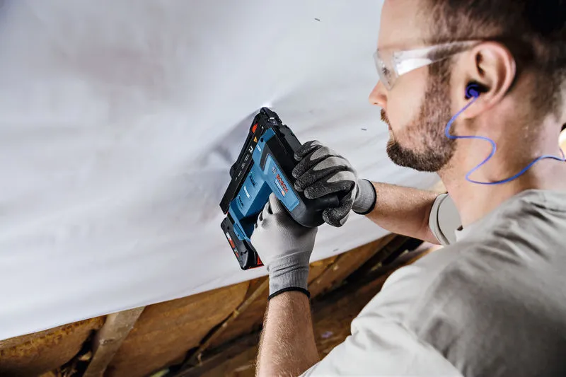 A person wearing safety equipment secures material to a ceiling using a cordless nail gun.