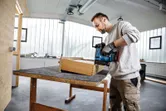 Person wearing safety equipment uses a cordless nail gun on a wooden box in a workshop.