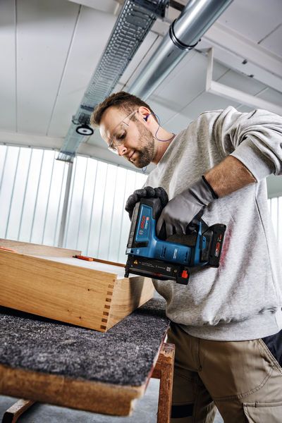 A person wearing safety equipment uses a cordless nail gun to fasten wooden joints.