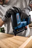 A person wearing safety equipment uses a cordless nail gun on a wooden surface.