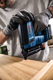 A person wearing safety equipment uses a cordless nail gun on a wooden surface.