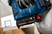 A person wearing safety equipment uses a cordless nail gun on a wooden plank.