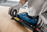 A person wearing safety equipment loads nails into a cordless nail gun on a wooden surface.