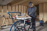 A person wearing safety equipment cuts wood on a table saw with dust extraction attached.