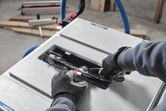 A person wearing safety equipment adjusts a table saw blade with a wrench.