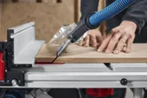 A person pushes wood through a table saw with dust extraction in a workshop.