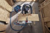 A person wearing safety equipment guides plywood through a table saw in a workshop.