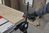 Person wearing safety equipment secures a wooden board on a table saw with a clamp.