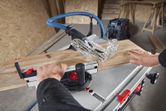A person guides a wooden board through a table saw in a workshop.