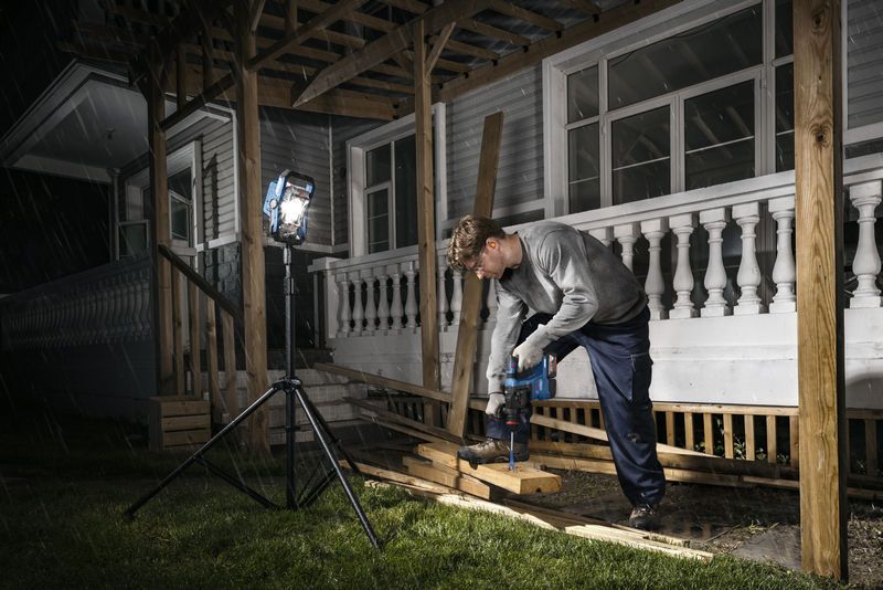 A person wearing safety equipment drills wood on a porch, illuminated by a cordless jobsite light.