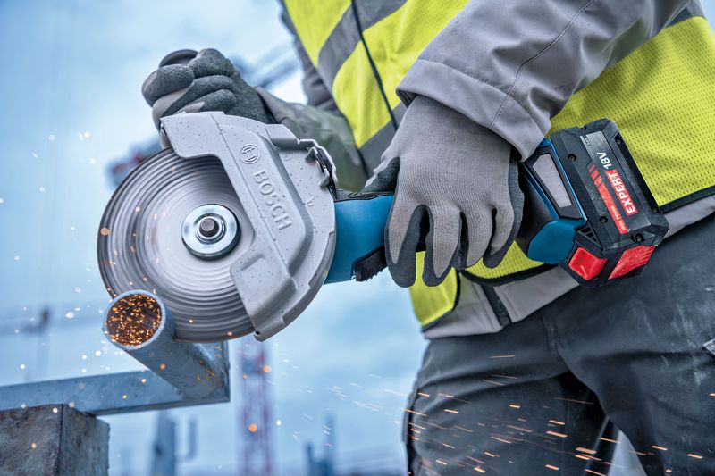 A person wearing safety equipment cuts a metal pipe with a cordless angle grinder.