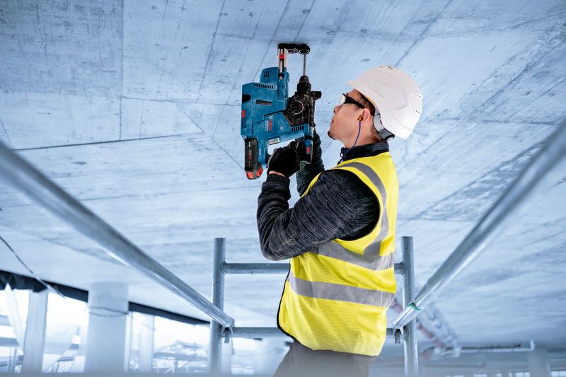 A person wearing safety equipment operates a rotary hammer drill on a concrete ceiling.