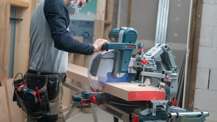 Object #200585317: 7557711_app_web_03 Person wearing safety equipment operates a miter saw to cut a wooden beam.