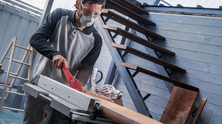 Object #200585338: 7557712_app_web_03 Person wearing safety equipment uses a table saw to cut wood indoors.