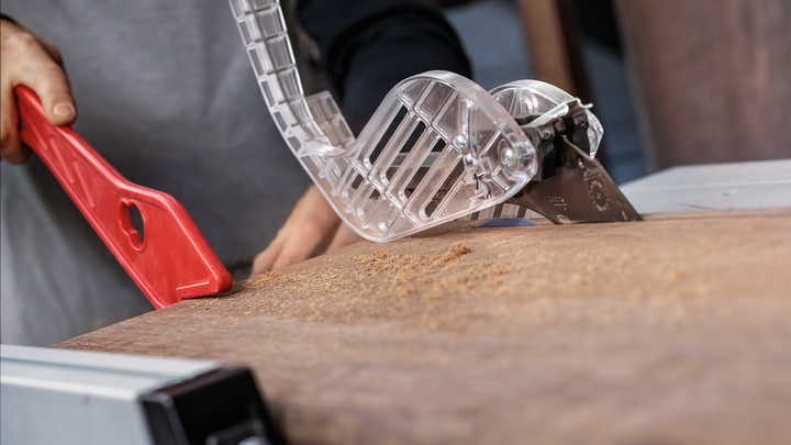 Object #200585342: 7557712_app_web_07 Person uses a push stick to guide wood through a table saw with safety guard.