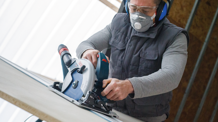 Object #200585533: 7557691_app_web_10 Person wearing safety equipment guides a circular saw along a board.