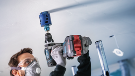 Person wearing safety equipment drills a large hole into a ceiling with a power tool.
