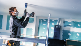 Person wearing safety equipment drills a hole into a ceiling at a construction site.