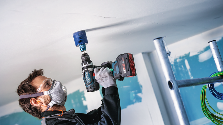 Person wearing safety equipment drills a circular hole into a ceiling on a construction site.