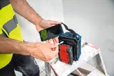 A person wearing safety equipment connects a smartphone to a portable Bluetooth speaker on a ladder.