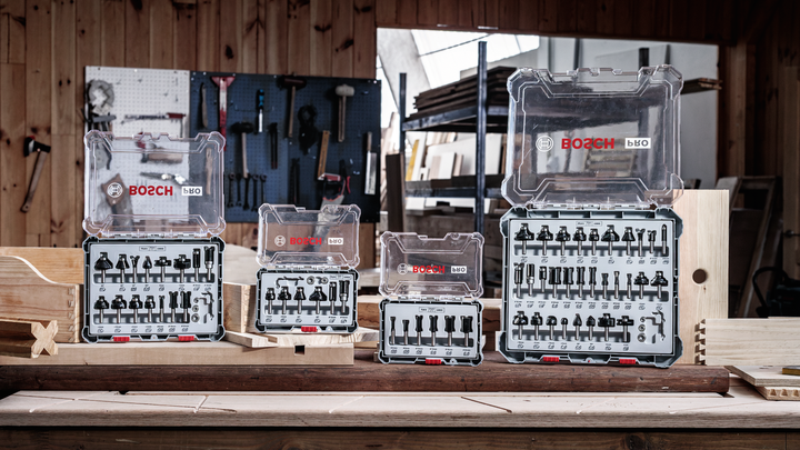 Sets of assorted drill and router bits arranged on a wooden workbench in a carpentry workshop.