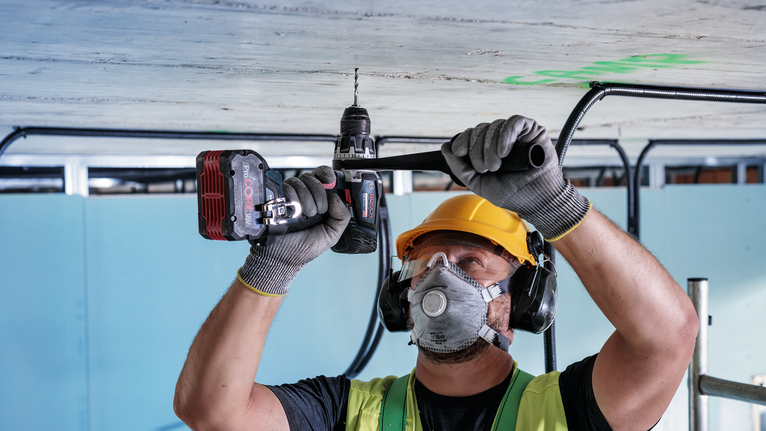 A person wearing safety equipment drills into a concrete ceiling with a cordless tool.