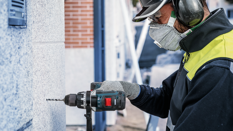 Person wearing safety equipment drills into a concrete wall.