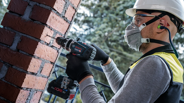 Person wearing safety equipment drills into a brick pillar outdoors.