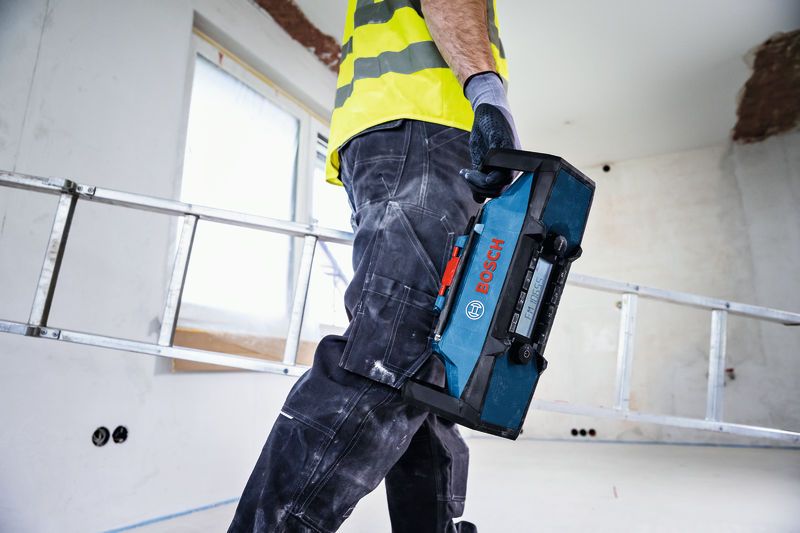 A person wearing safety equipment carries a blue jobsite radio in a construction area.