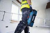 A person wearing safety equipment carries a blue jobsite radio in a construction area.
