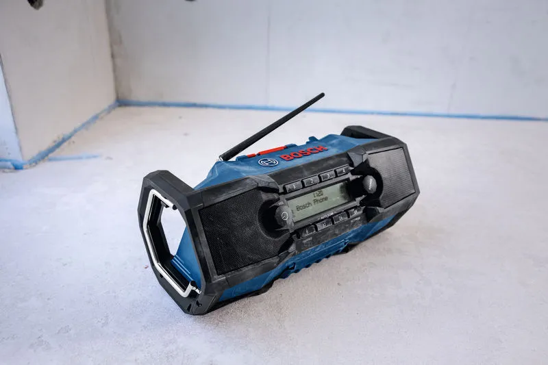 Portable jobsite radio with display and two speakers sits on a concrete floor.