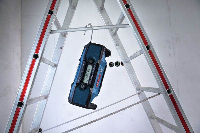 Job site radio hanging from a metal hook on a ladder in a construction area.