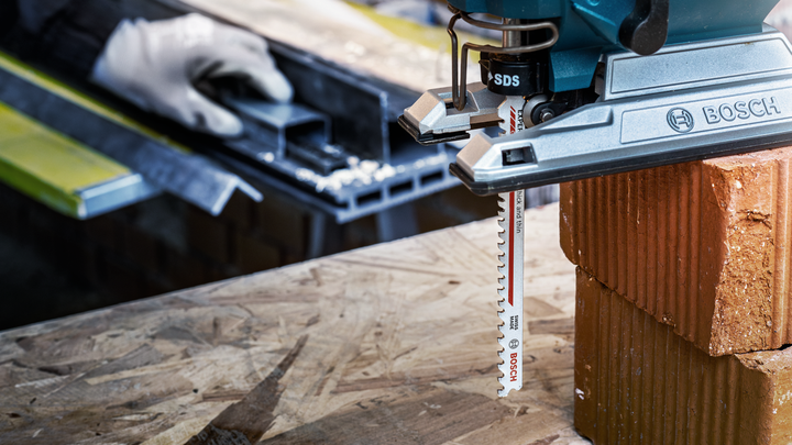 Person wearing safety equipment uses a jigsaw to cut a brick on a wooden surface.