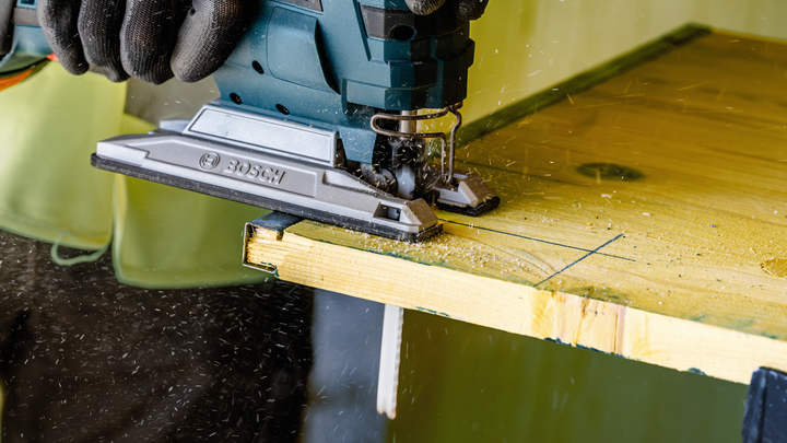 A person wearing safety equipment uses a jigsaw to cut a wooden board.