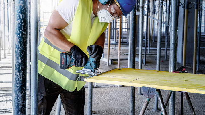 Person wearing safety equipment uses a jigsaw to cut wood on a construction site.