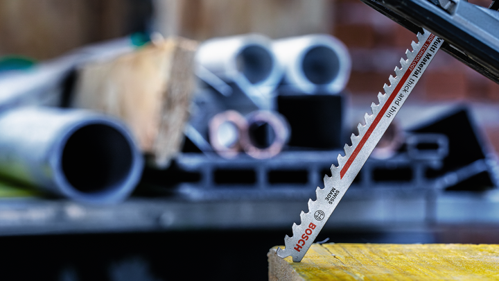 Saw blade cutting through wood with metal pipes and construction materials in the background.