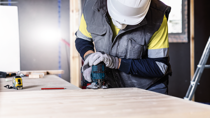 Person wearing safety equipment uses a power tool to cut wood at a workbench.