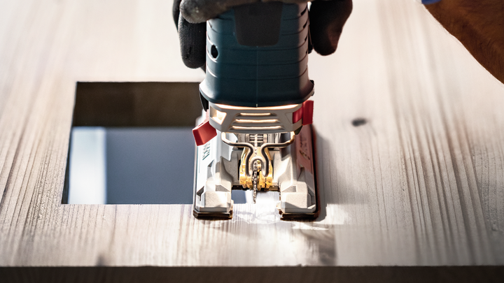 Person wearing safety equipment uses a jigsaw to cut a square opening in wood.