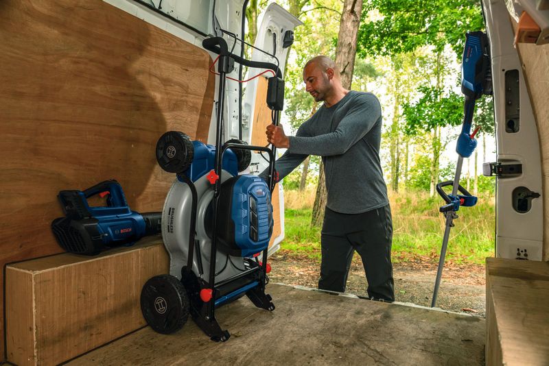 A person loads a blue lawn mower into a work van parked next to a wooded area.