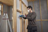 A person wearing safety equipment uses a multifunction tool to cut drywall.